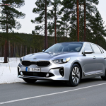 A sophisticated silver Kia K8 Hybrid sedan is cruising smoothly on a well-paved Swedish rural road during a crisp winter day. The car's elegant lines are visible against a backdrop of snow-dusted pine trees and a clear sky. Emphasize the vehicle's stable presence and the serene atmosphere of the northern landscape. Ultra-realistic, cinematic, serene.